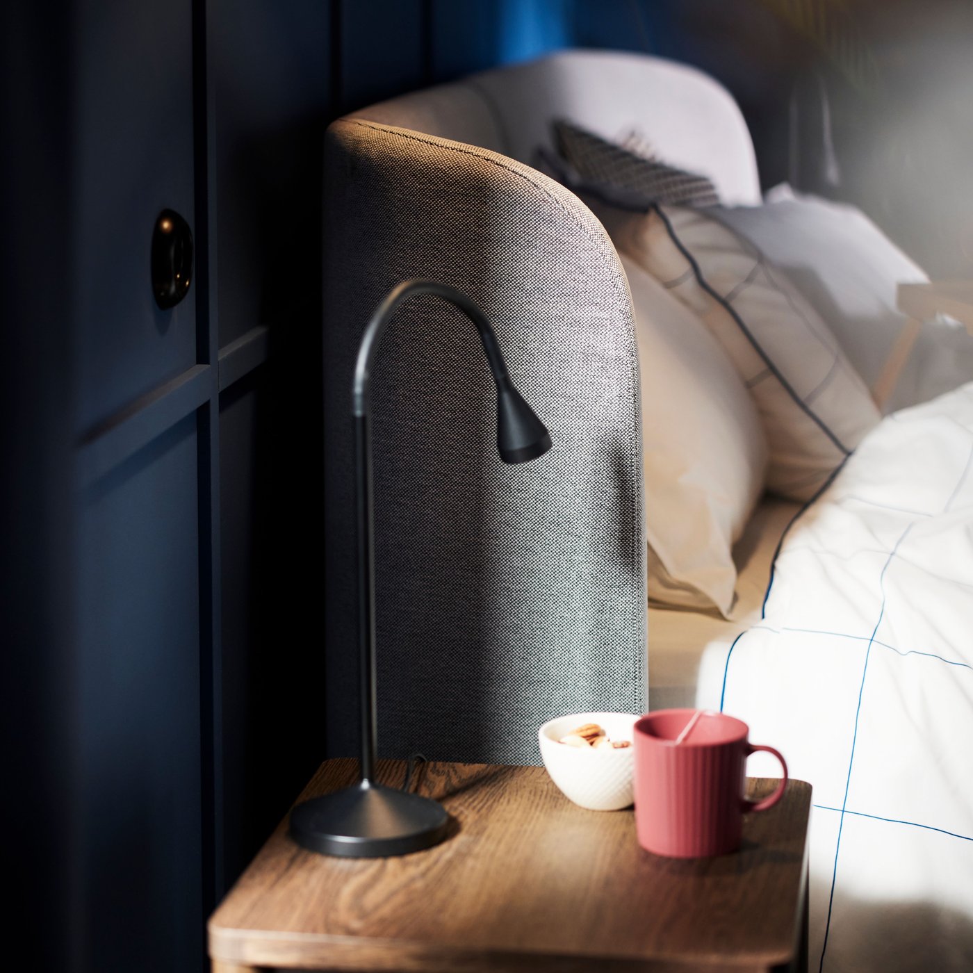 Huvudgaveln på en TUFJORD klädd säng med ett TONSTAD sängbord bredvid, ovanpå står en svart NÄVLINGE LED-arbetslampa.