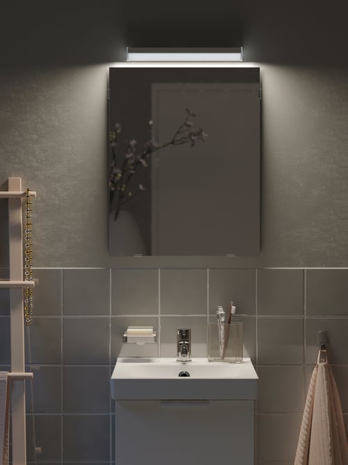 Baie cu o oglindă dreptunghiulară din sticlă iluminată cu un sistem de iluminare LED amplasat lângă un suport pentru prosoape.