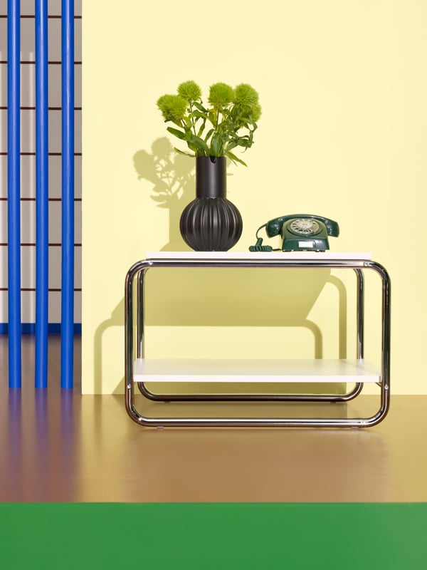 A white BAGGBODA side table stands in front of a light yellow wall. The table holds a black vase and an old phone.