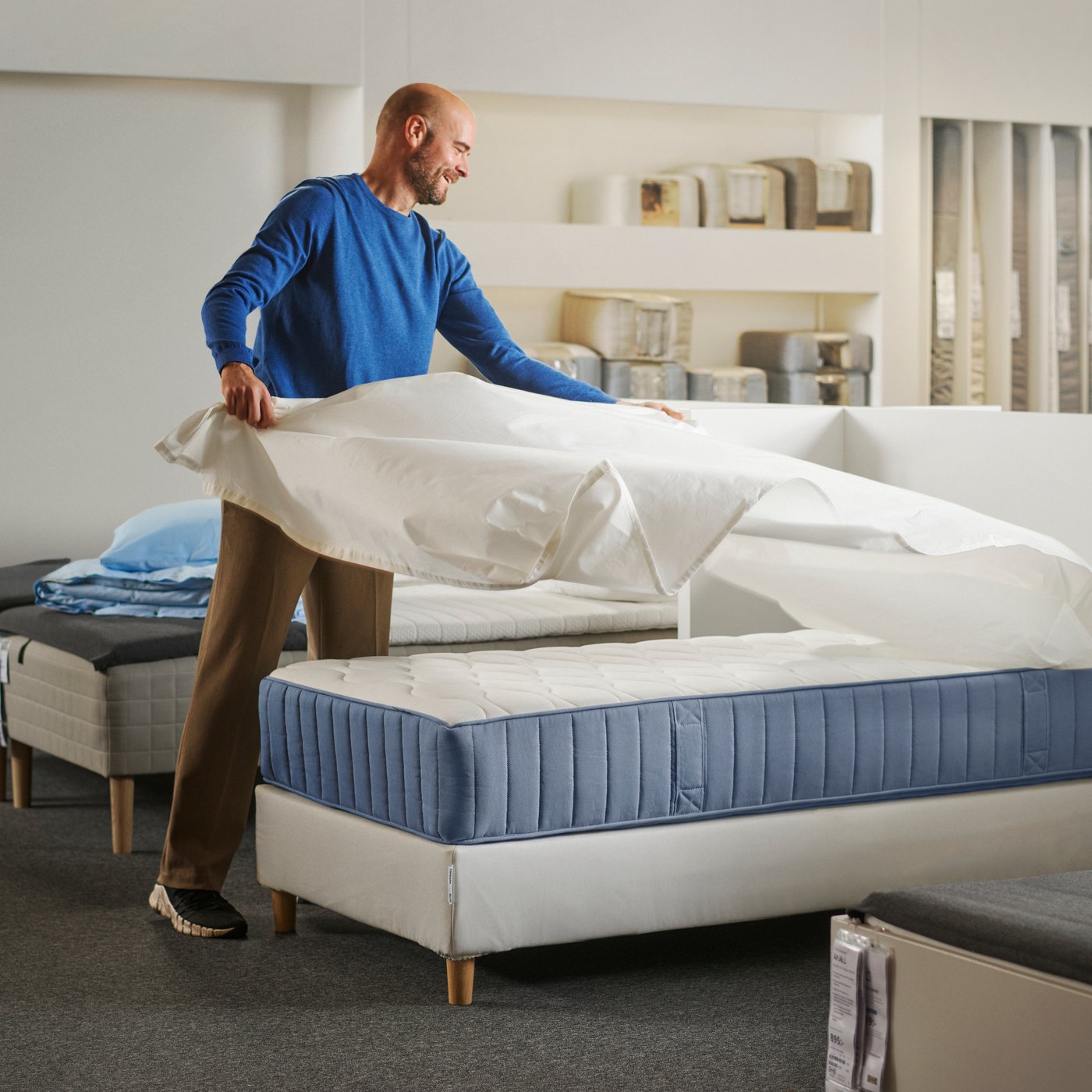 En person som bäddar en VÅGSTRANDA resårmadrass med pocketfjädrar med ett lakan i en butiksmiljö.