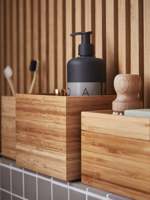 Three bamboo boxes placed on a tiled surface with a soap dispenser, two toothbrushes, and a container with wooden cap.