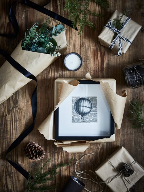 Holzoberfläche mit einem schwarzen SANNAHED Rahmen, der gerade zwischen einer BEHJÄRTAD Duftkerze, Dekorationen und eingepackten Geschenken ausgepackt wird.