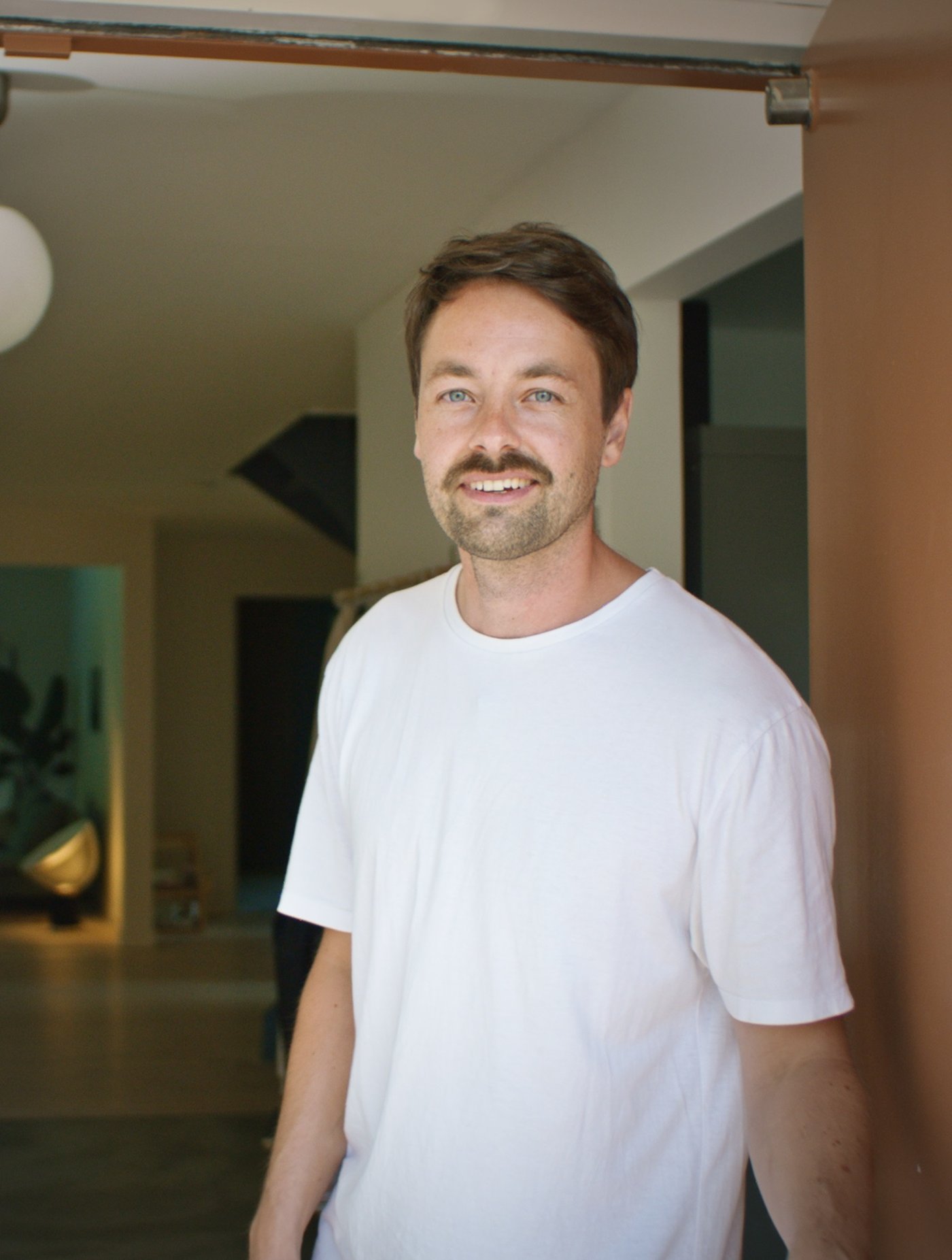 Muškarac smeđe kose u beloj majici kratkih rukava stoji na ulazu u kuću.