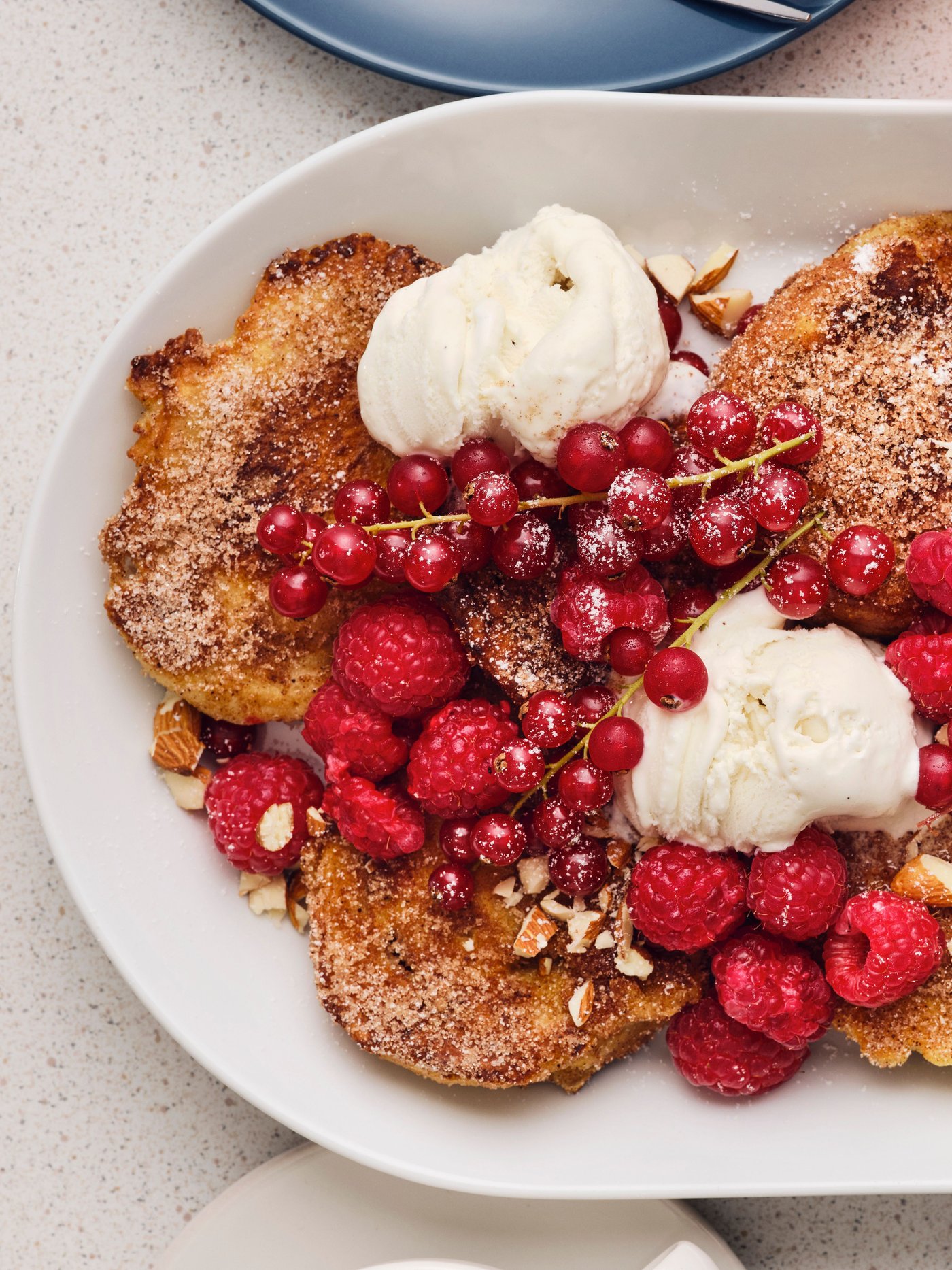 Auf einem weissen, ovalen Teller liegen mehrere Stücke French Toast mit Eis, frischen Himbeeren, roten Johannisbeeren und Mandeln.