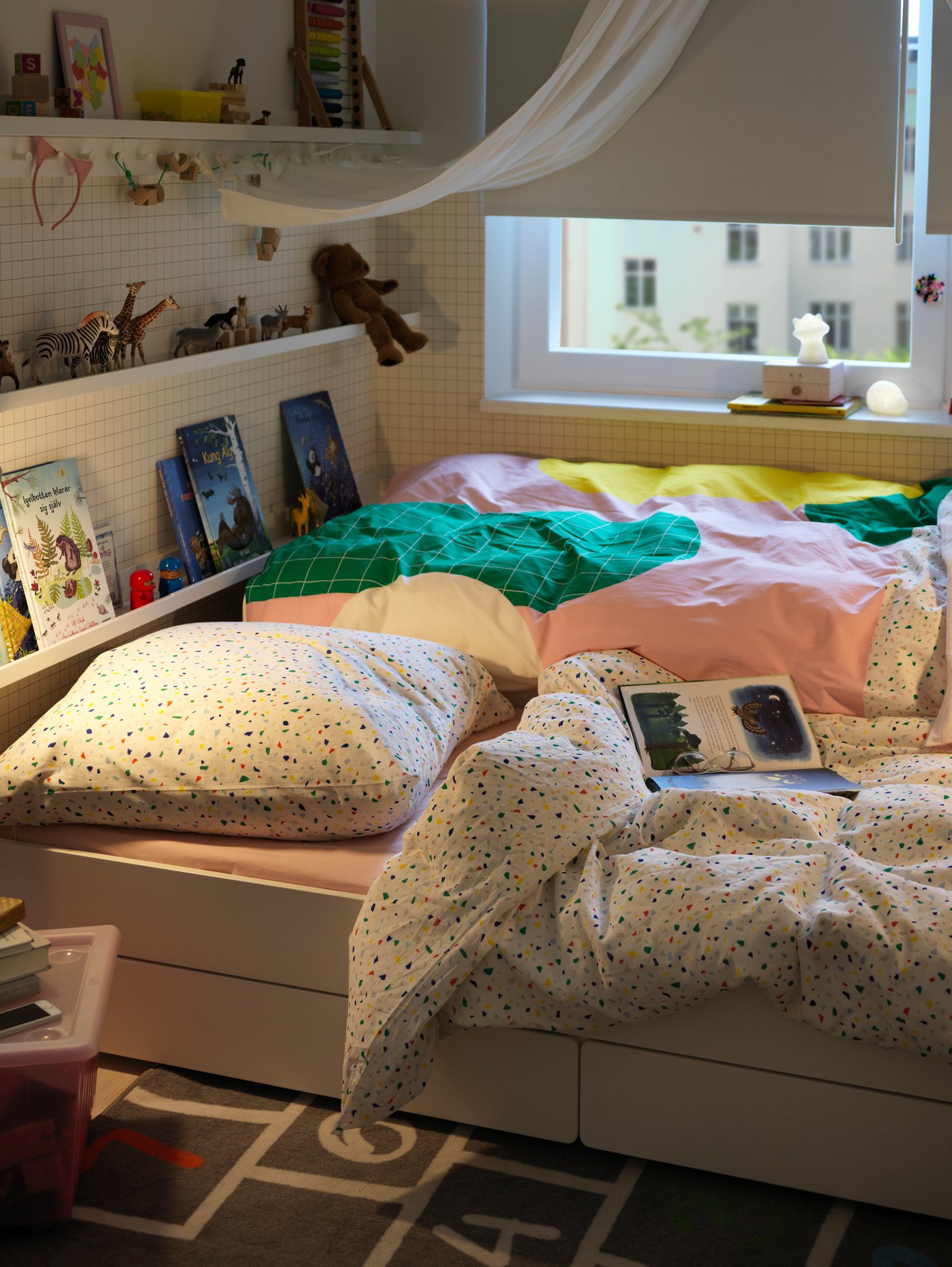 Dormitorio infantil con estructura de cama con debajo de la cama, estor, fundas nórdicas y de almohada, libros, juguetes, alfombra, ventana y cortinas.