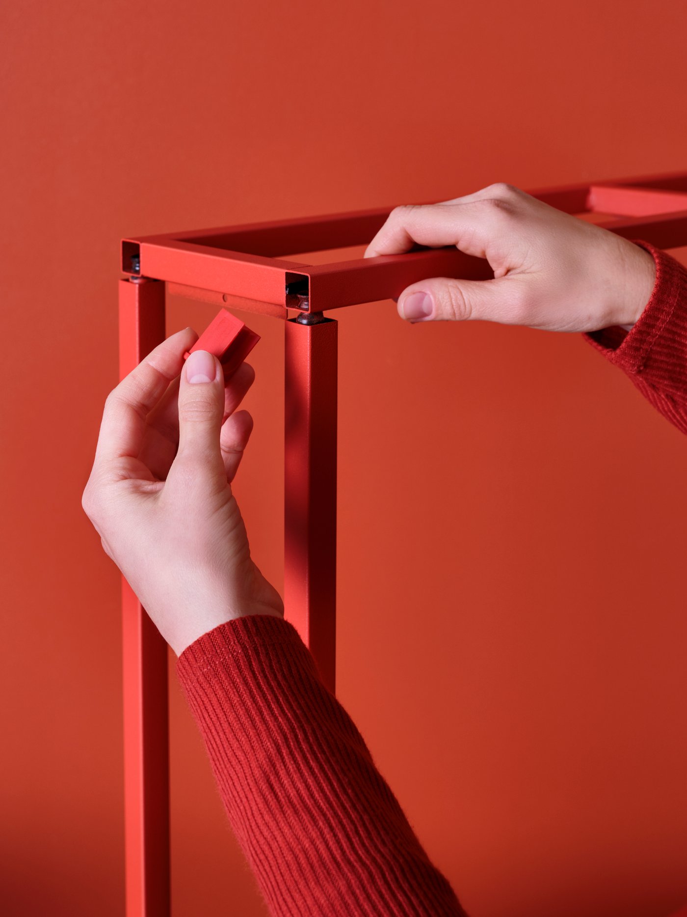 صورة عن قرب ليد تثبت وتدًا خشبيًا داخل إطارات ENHET معدنية حمراء، أمام خلفية باللون الأحمر.