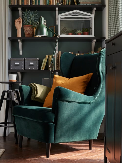 Un fauteuil à accoudoirs STRANDMON vert foncé devant des tablettes avec un jeté ODDRUN vert et un coussin avec housse VIGDIS jaune foncé.