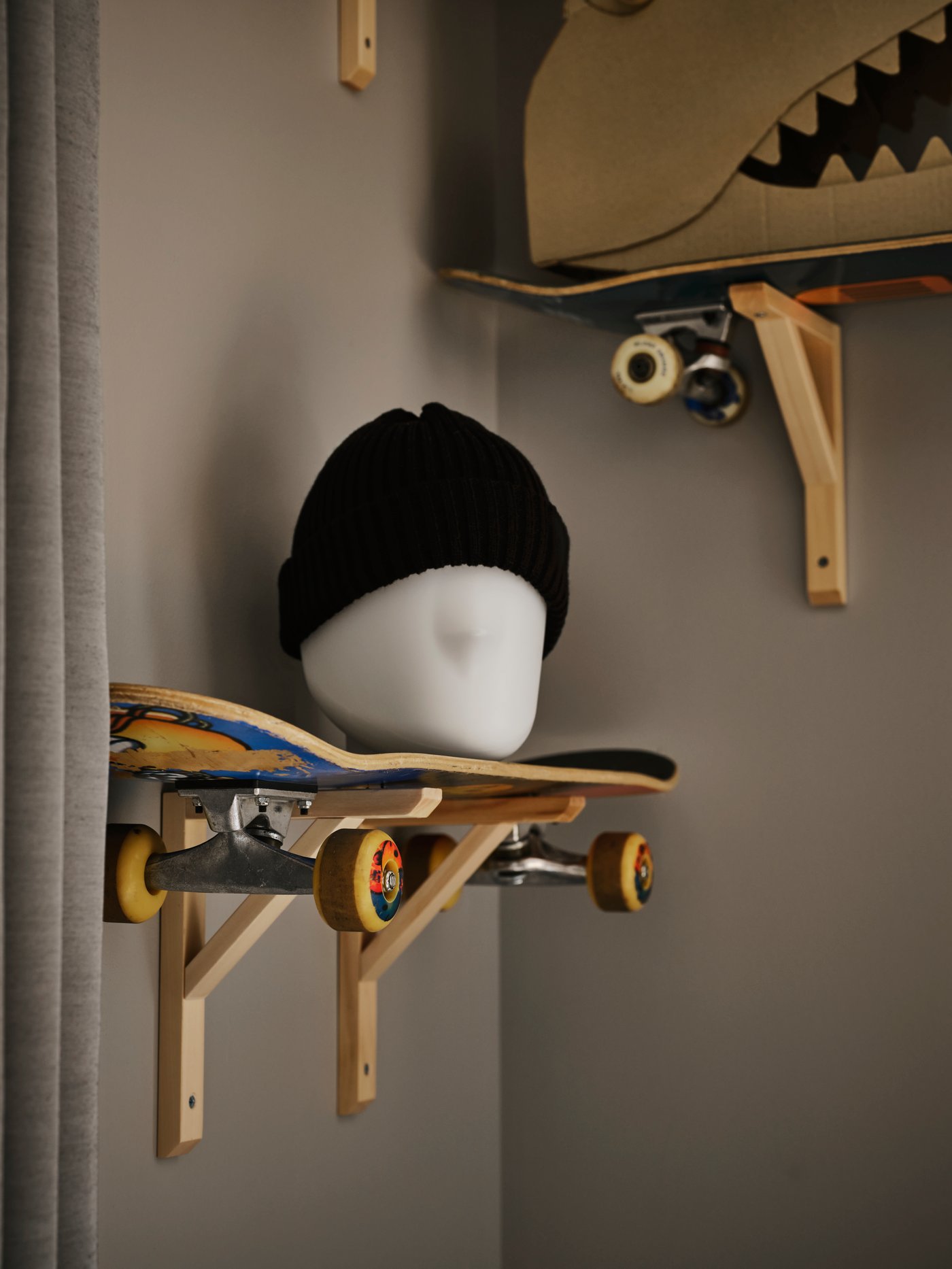 En svart ullmössa placerad ovanpå LED-bordslampan ISKÄRNA ovanpå en hylla som består av en skateboard, som hänger i ett litet tonårsrum.