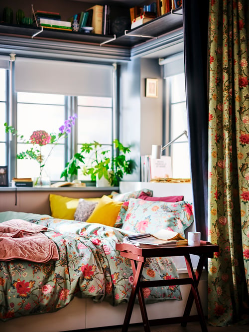 A MARYD tray table next to a bed with a NÄSSELKLOCKA quilt cover in a windowed recess with plants on the windowsill.