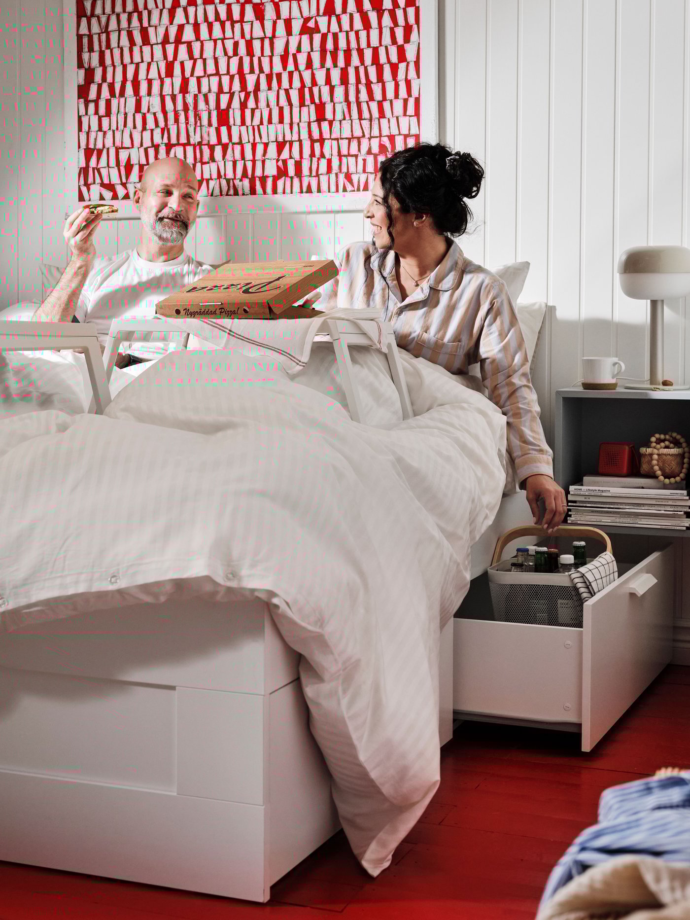 BRIMNES säng med vitt bäddset och uppfällda brickbord, pizzakartong och mat, med öppen låda fylld med förvaringskorg och småsaker samt sängbord med lampa och kopp.