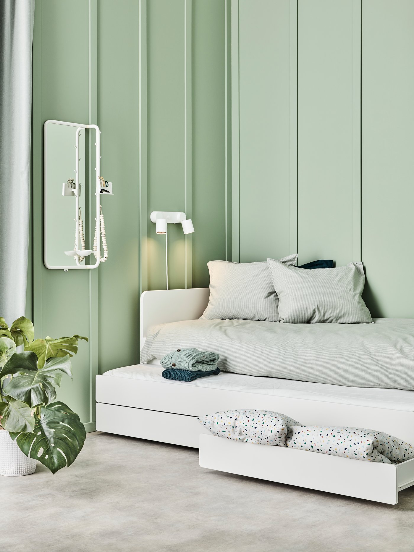 Dormitorio con estructura de cama con cama inferior y cajones, fundas nórdicas, fundas de almohada, lámpara de pared, espejo, alfombra, maceta y ropa.