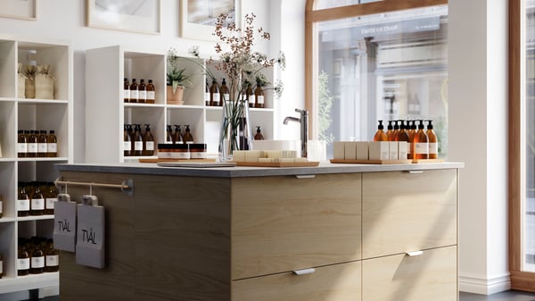 Un negozio ampio e luminoso e un'isola cucina con frontali per cassetti ASKERSUND effetto frassino chiaro, con lampade a sospensione SÖDÅKRA.