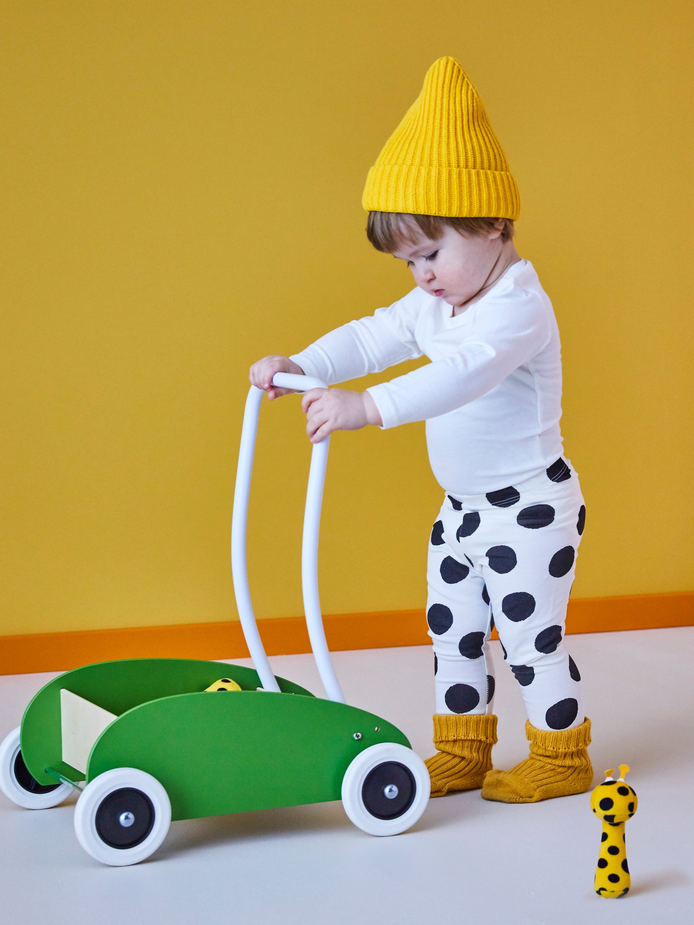 A child wearing a yellow hat and black and white spotty trousers pushes a green MULA toddle truck past a soft toy.