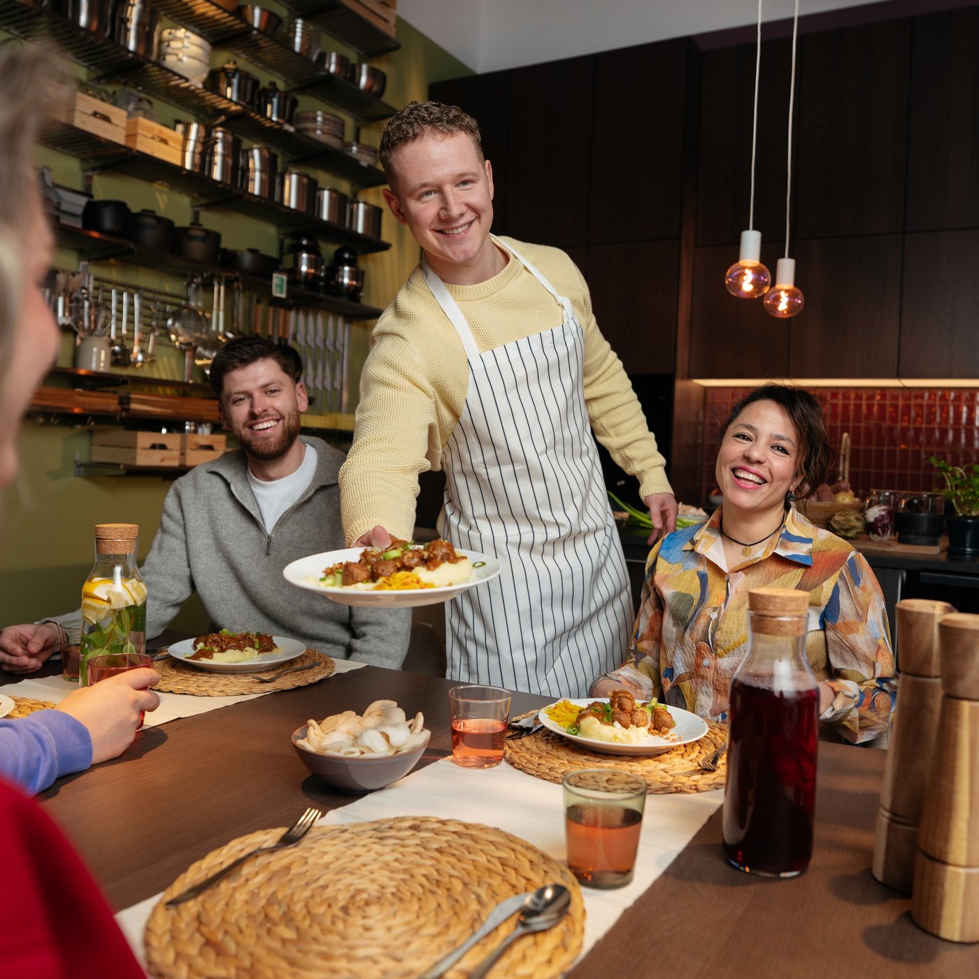 A man wearing an apron is holding a plate of food. Around him, people are sitting and smiling as they look at his dish.