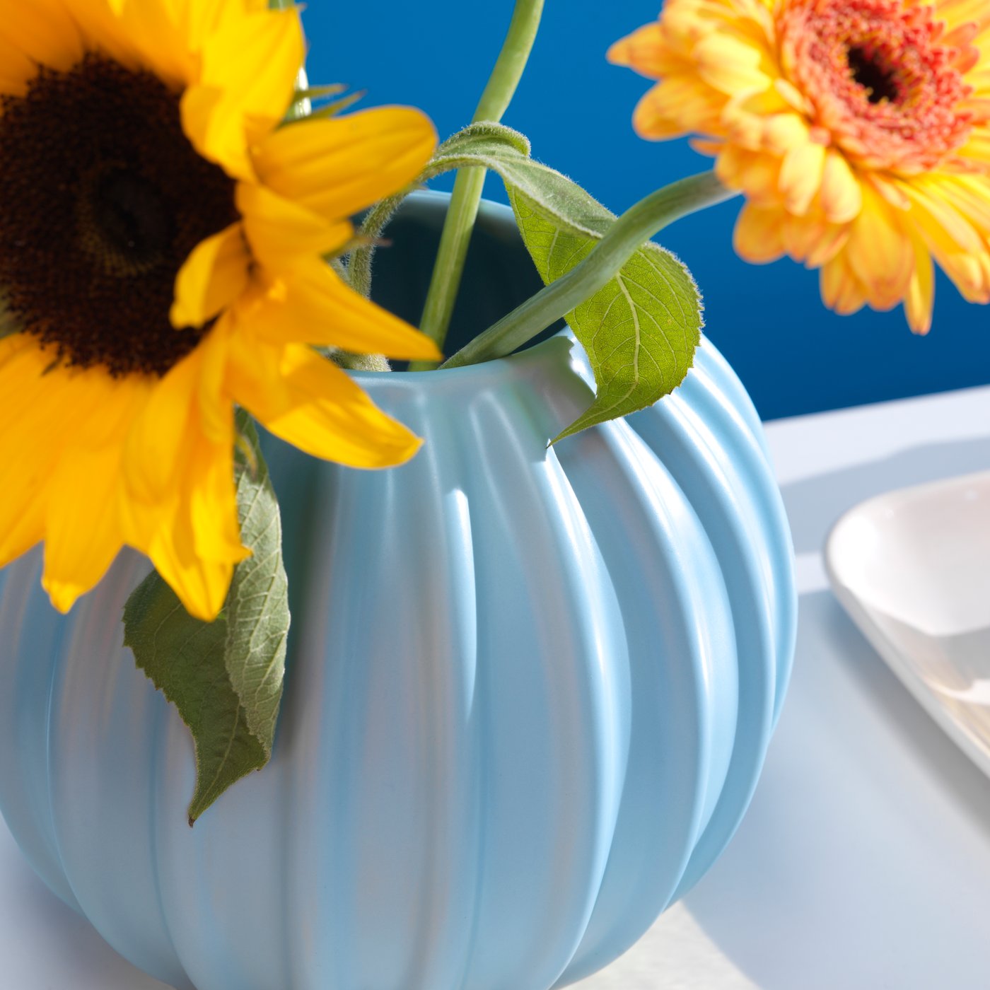 Los girasoles amarillos están en un florero SKOGSTUNDRA azul claro, que está sobre una mesa blanca contra una pared azul.
