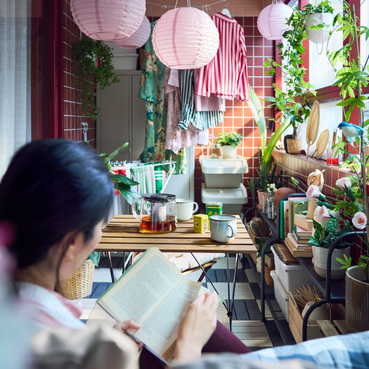 A lively balcony with green plants and pink lamps, featuring a black and light brown wooden TÄRNÖ outdoor table and tea mugs.