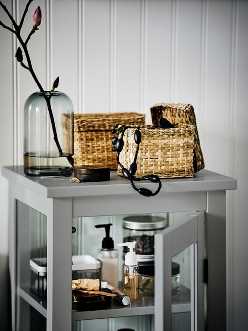Two LURPASSA seagrass boxes with lids and a grey DRÖMSK glass vase on a grey HEMNES high cabinet with glass door.