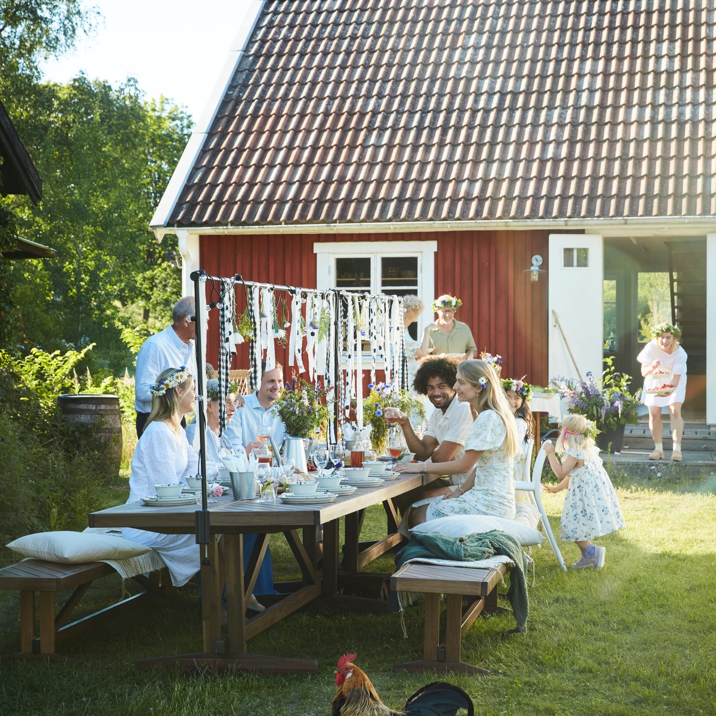 Family and friends gather around a NORRMANSÖ table in the garden, sharing food and enjoying midsummer eve.