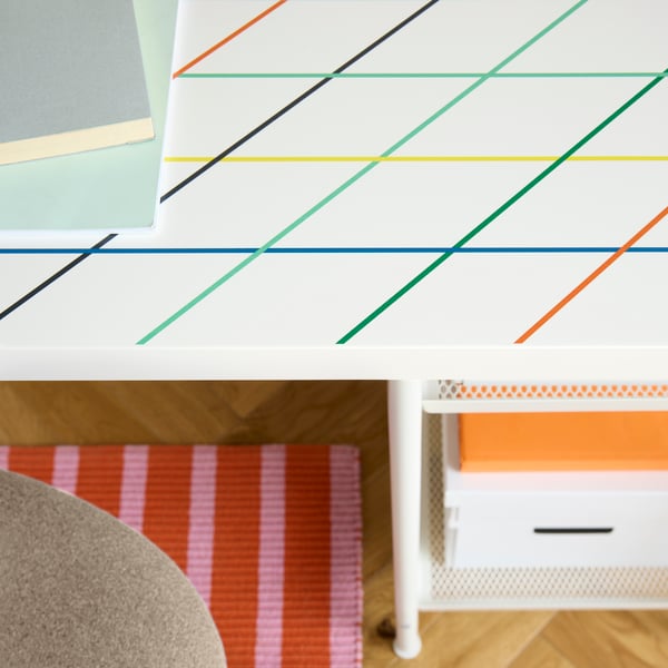 A close-up of a multicoloured LAGKAPTEN tabletop with white SPÄND legs and a striped rug underneath the chair.