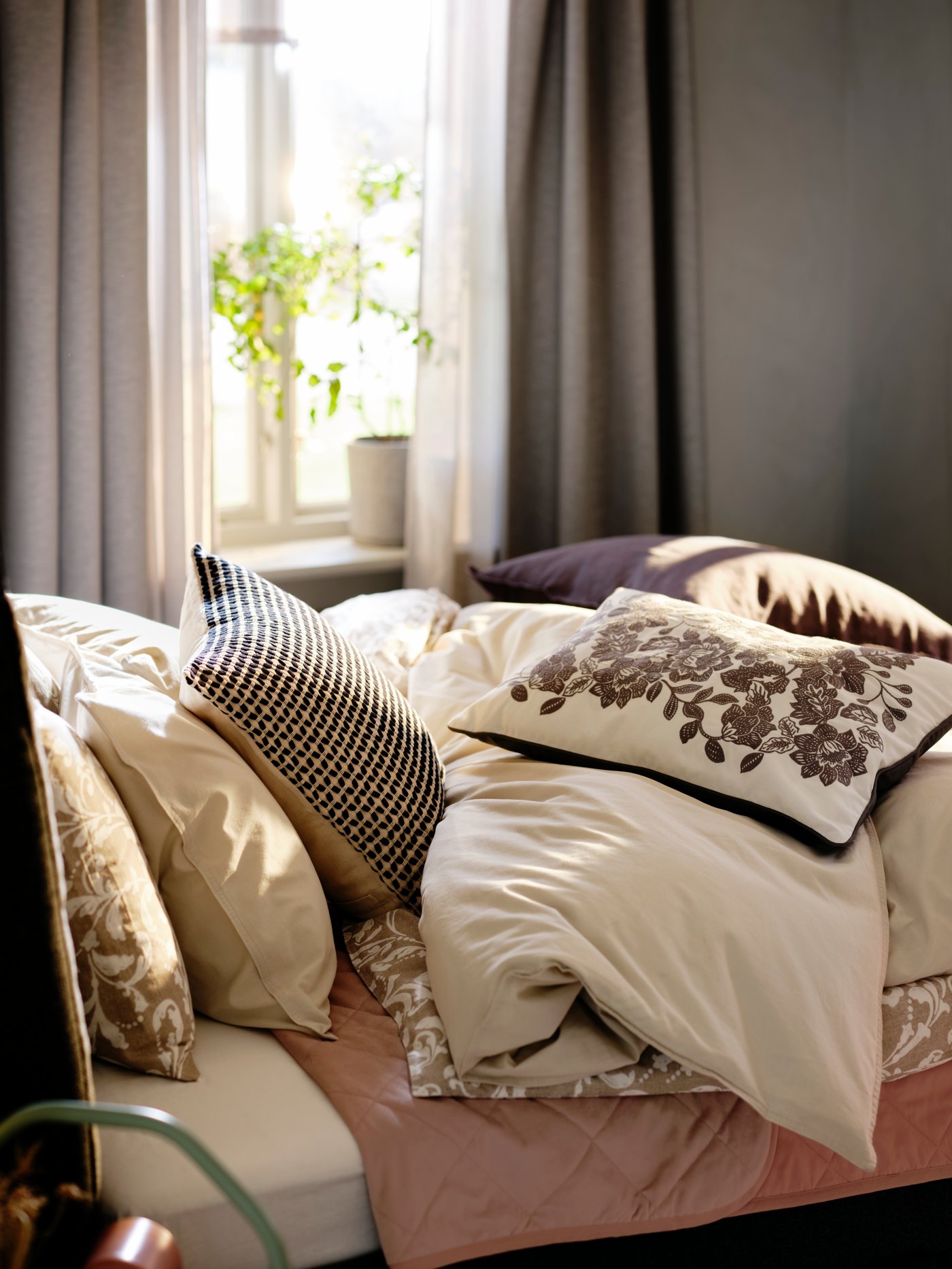 Parte superior de una cama cubierta con una mezcla de textiles de colores neutros, como ÄNGSLILJA, SKUGGNÄVA y VÅRBRACKA.