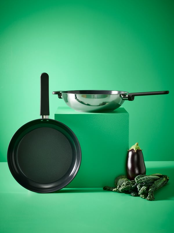 A MIDDAGSMAT wok in stainless steel placed on green pedestal next to a MIDDAGSMAT frying pan, an aubergine and kale.