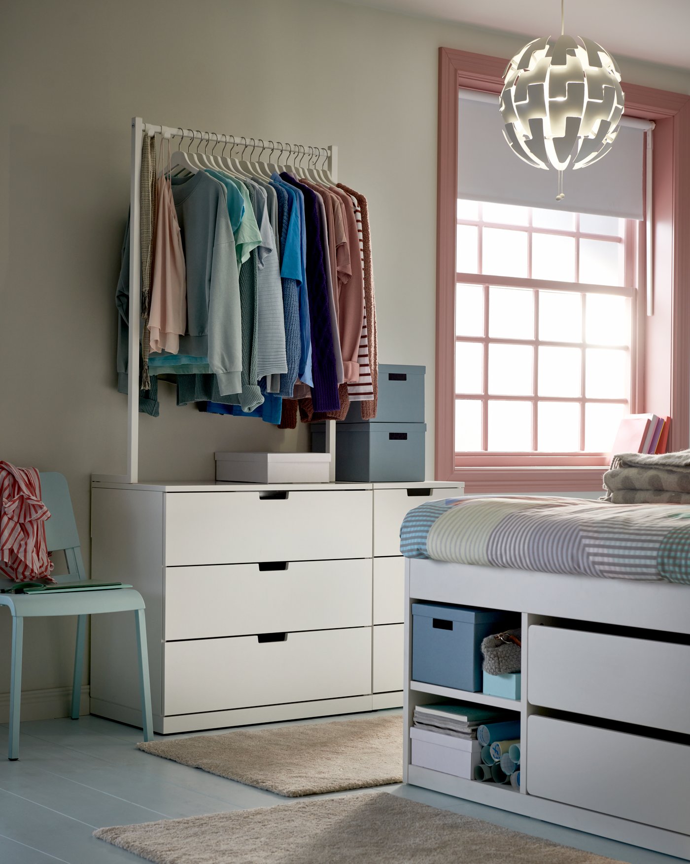 Bedroom with white dresser with clothes rail on top, colorful shirts hanging, white NORDLI bed with storage drawers, blue chair, pink window frame and round white ceiling lamp.
