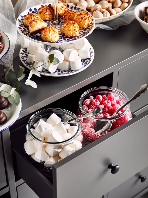Part of a gray BRYGGJA chest of drawers with glass jars and plates filled with sweets on top and inside a drawer.