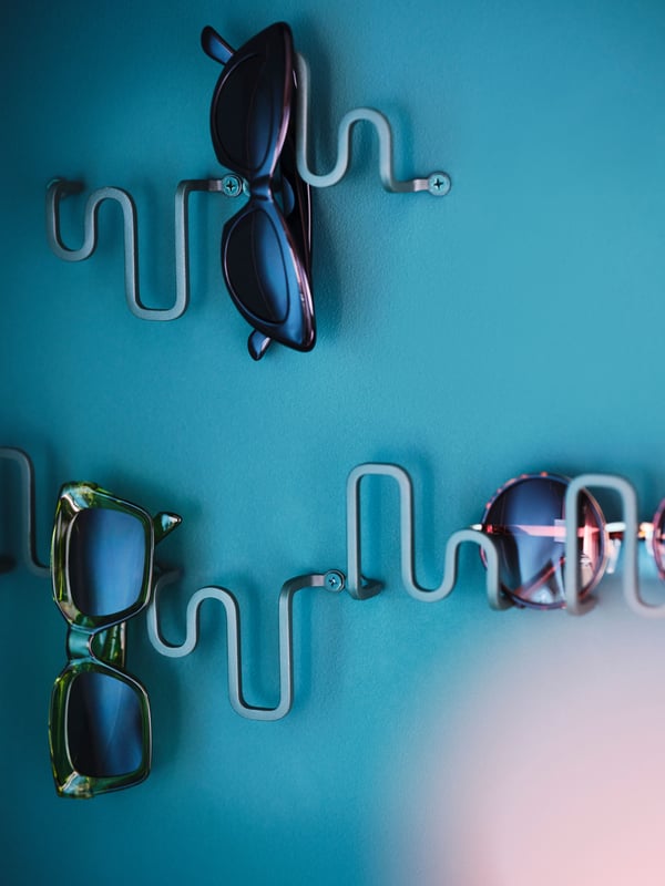 A few dark grey-green JORDMÅNEN hooks on a wall beside each other holding sunglasses at different angles.