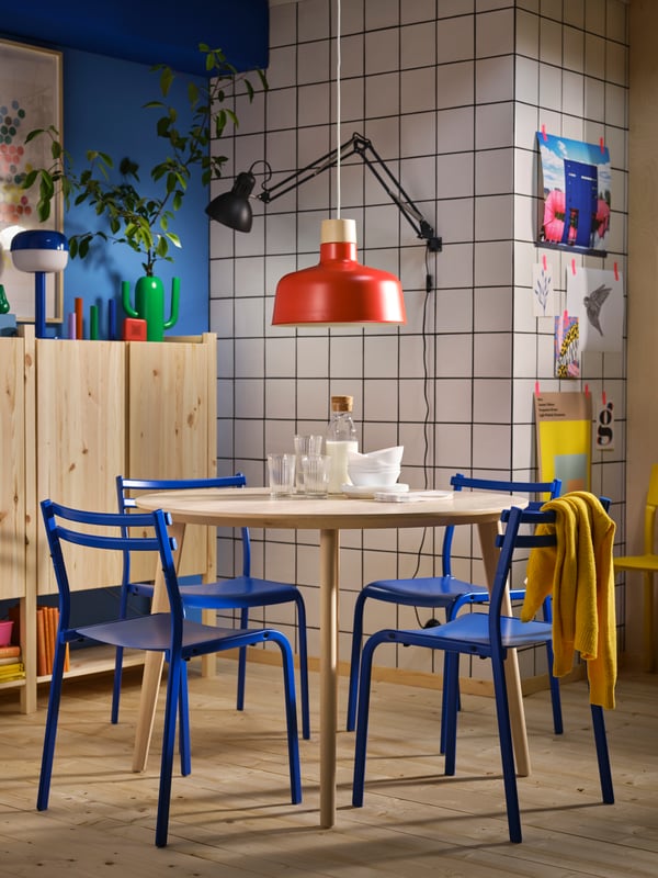 A bright dining area with blue GENESÖN chairs round a DOCKSTA table under a red BUNKEFLO pendant, and IVAR cabinets nearby.