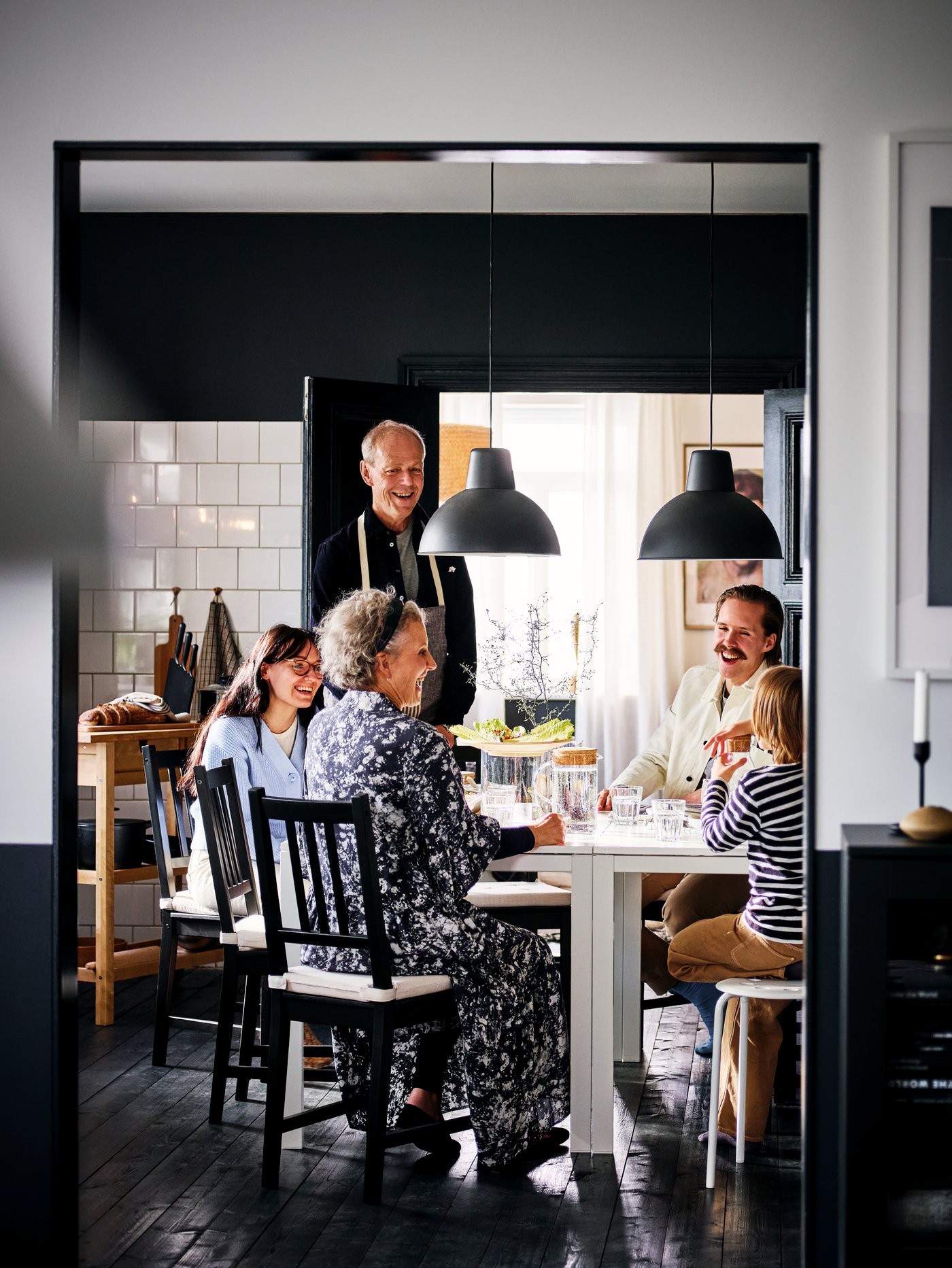 Comedor con varias personas sentadas alrededor de una mesa rectangular, en sillas marrones, debajo de 2 lámparas de techo negras.