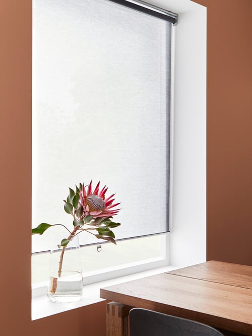 Window outfitted with a grey roller blind, a flower in a white vase on the windowsill, and a rectangular table to the side.