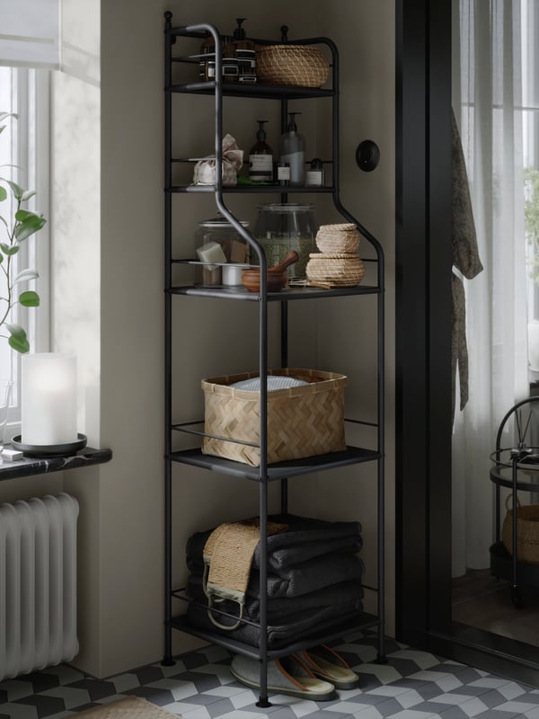 A black FRÖSJÖN shelving unit stands in a bathroom corner. It has five open shelves filled with baskets and spa essentials. 