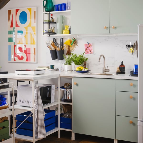 A kitchen with storage with ENHET doors in pale green-grey, ENHET wall frame with shelves in white and a colourful poster.