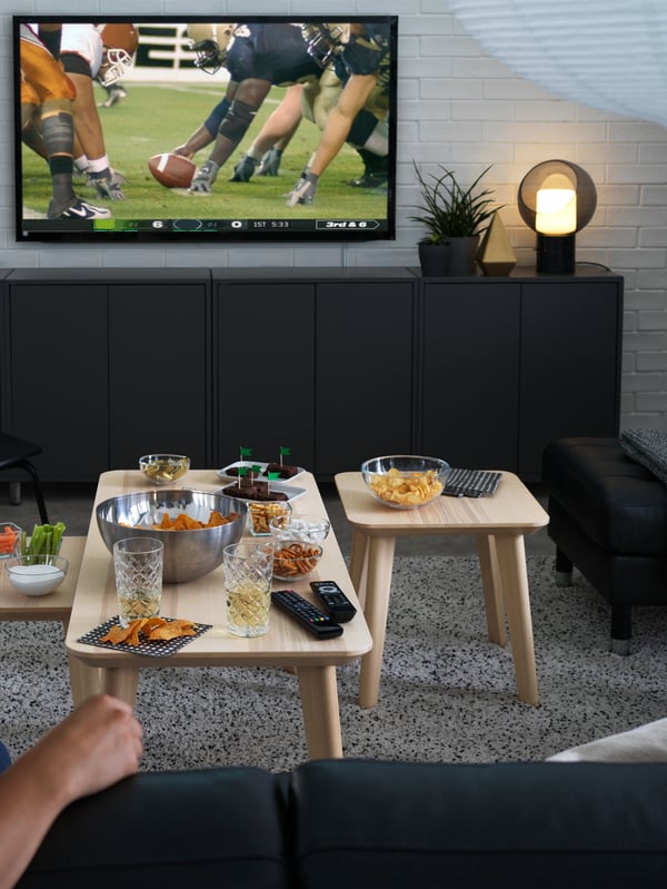 A side table and coffee table with bowls, glasses, and remotes placed in front of a TV, with a table lamp to the side.