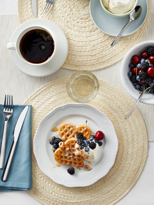 A table with four SLUTEN table mats on it. They have blue bowls and plates of food, cups of coffee and cutlery on them.