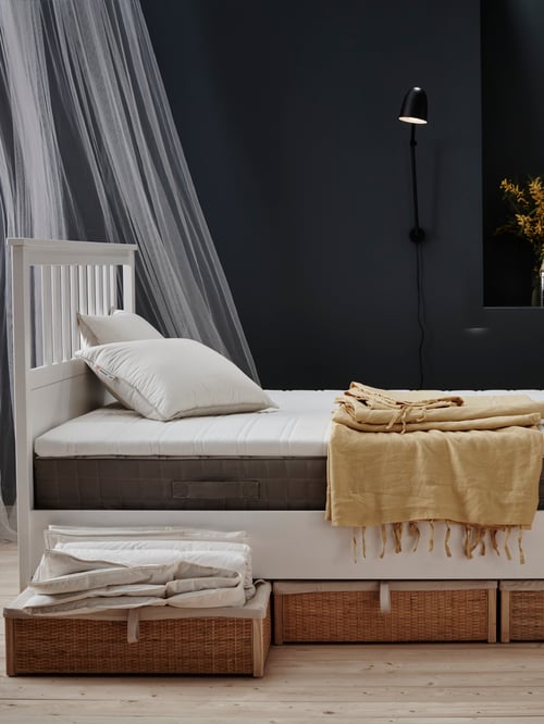 Bedroom with white wooden bed frame, mattress pad, sprung mattress, storage boxes, pillow, curtains, work lamp, throw, duvet.