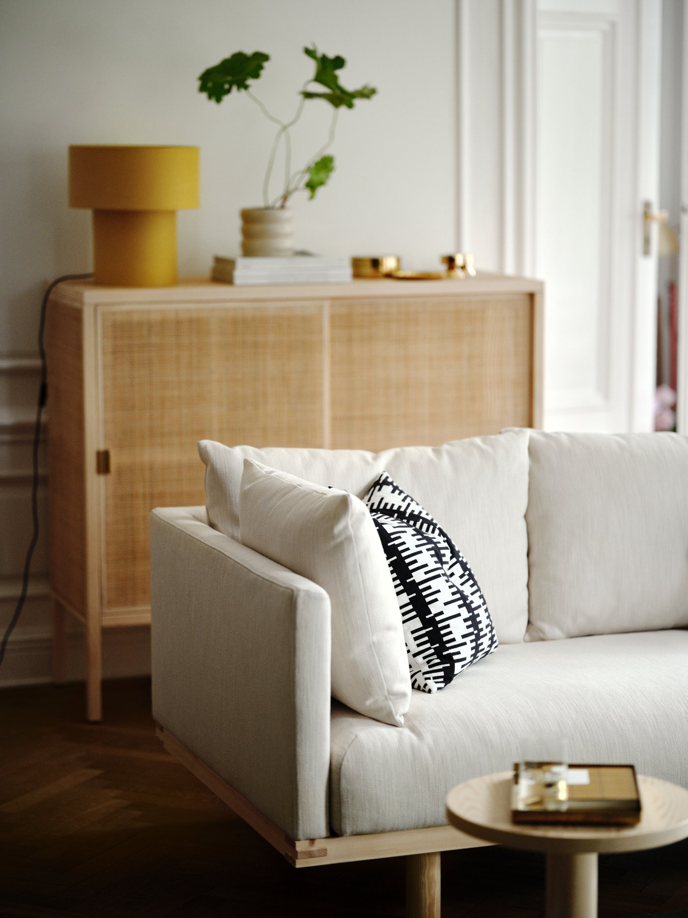 Een woonkamer met een beige bank met en een offwhite/zwarte STOCKHOLM 2025 kussenhoes en daarachter een houten kast met gele tafellamp.