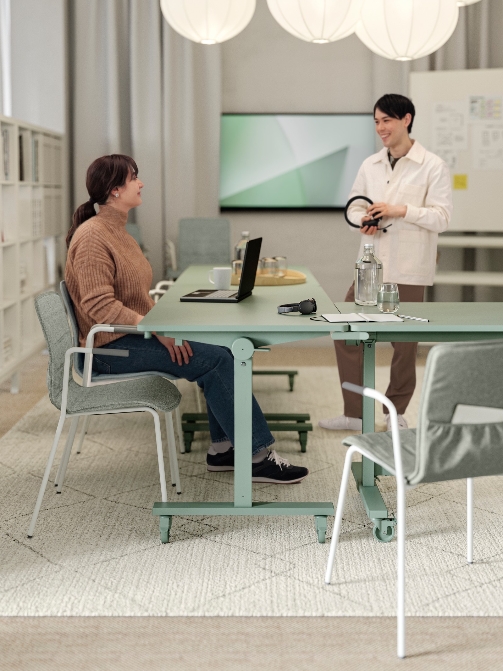 A woman and a man in a meeting area with three green MITTZON foldable tables with castors, a white rug and white round lamps.