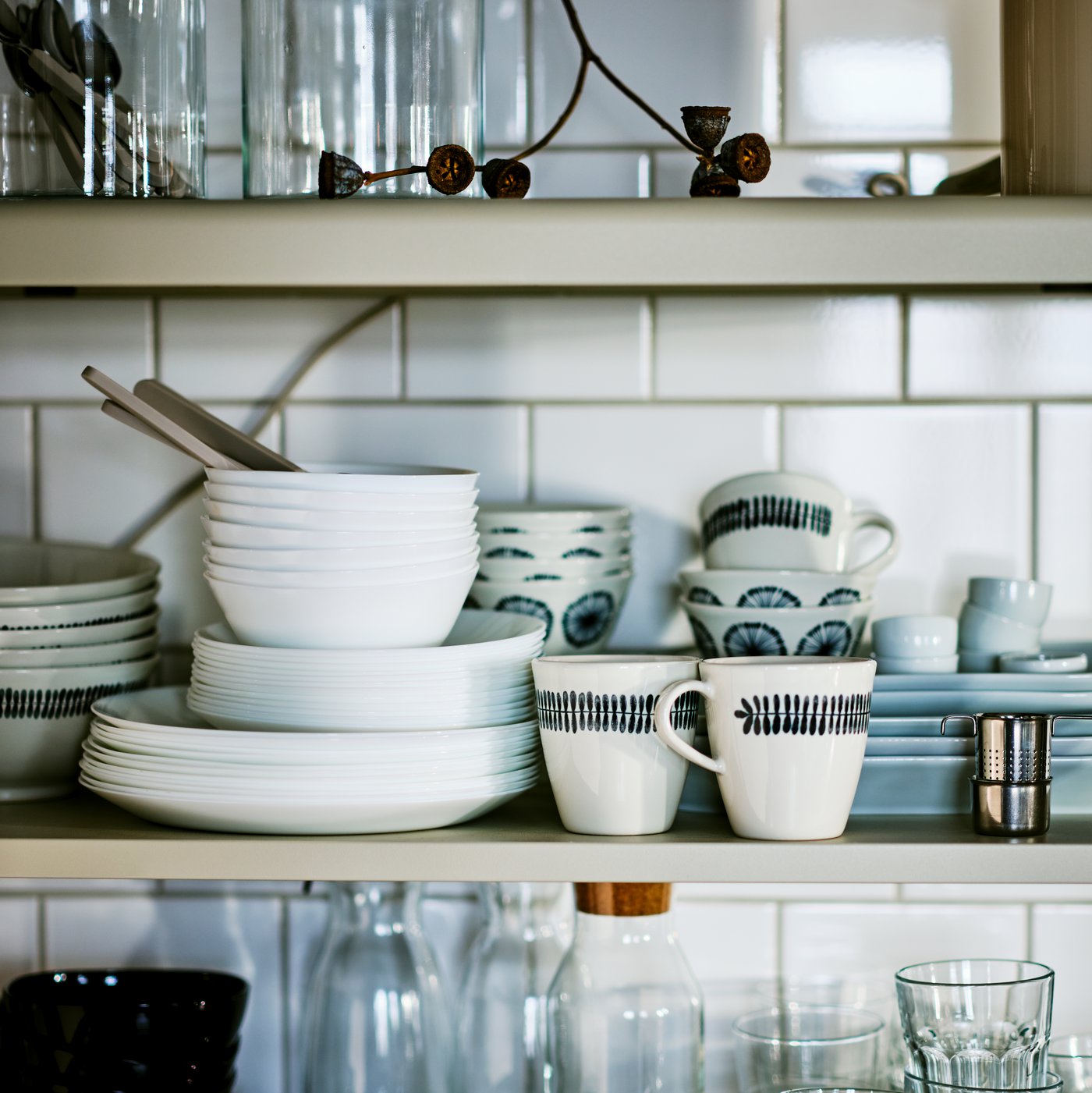 Shelves mounted on a white-tiled kitchen wall, holding stacks of white OFTAST bowls and plates, POKAL glasses, and more.