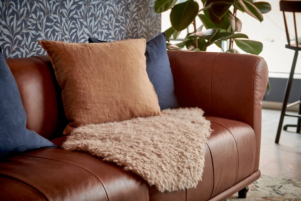 En VISKAFORS brun lädersoffa med blå och beiga kuddar, beige pläd, bladmönstrad tapet och grön växt vid fönster.