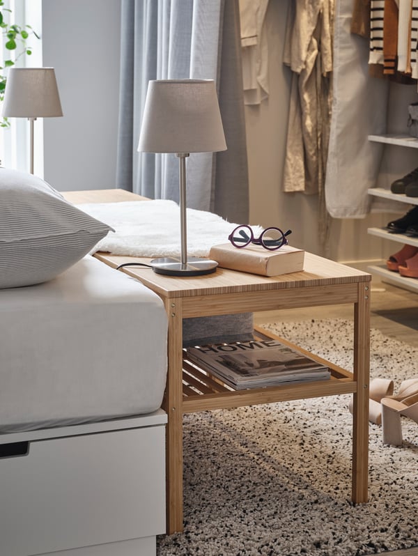 A bamboo NORDKISA bench stands beside a bed and is used as a bedside table, with table lamps, glasses and a book on top.