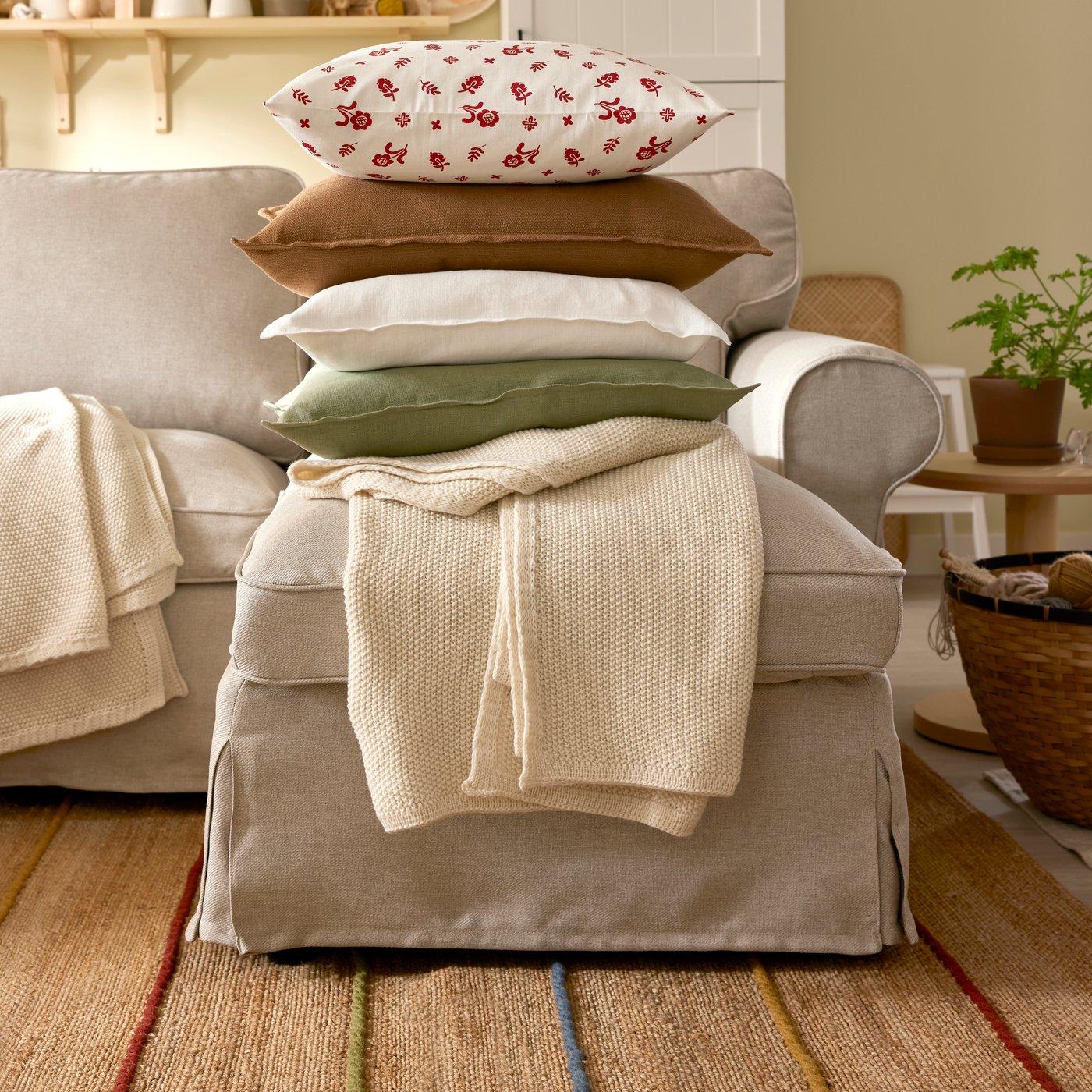 A pile of four cushions is on top of an off-white HUMLEMOTT throw on a light beige EKTORP 3-seat sofa with chaise longue.