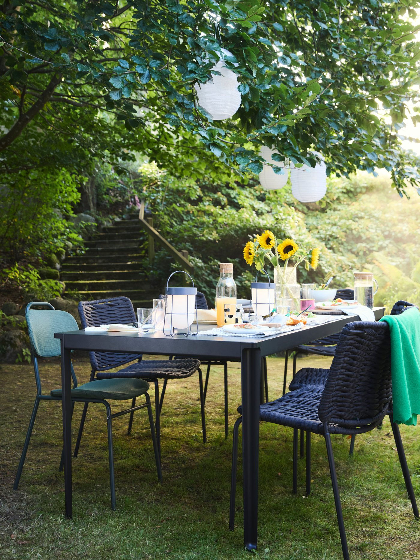 나뭇가지에 장식 조명이 매달려 있는 정원의 나무 아래에 TEGELÖN 테겔뢴 테이블이 놓여있는 야외 공간의 모습.