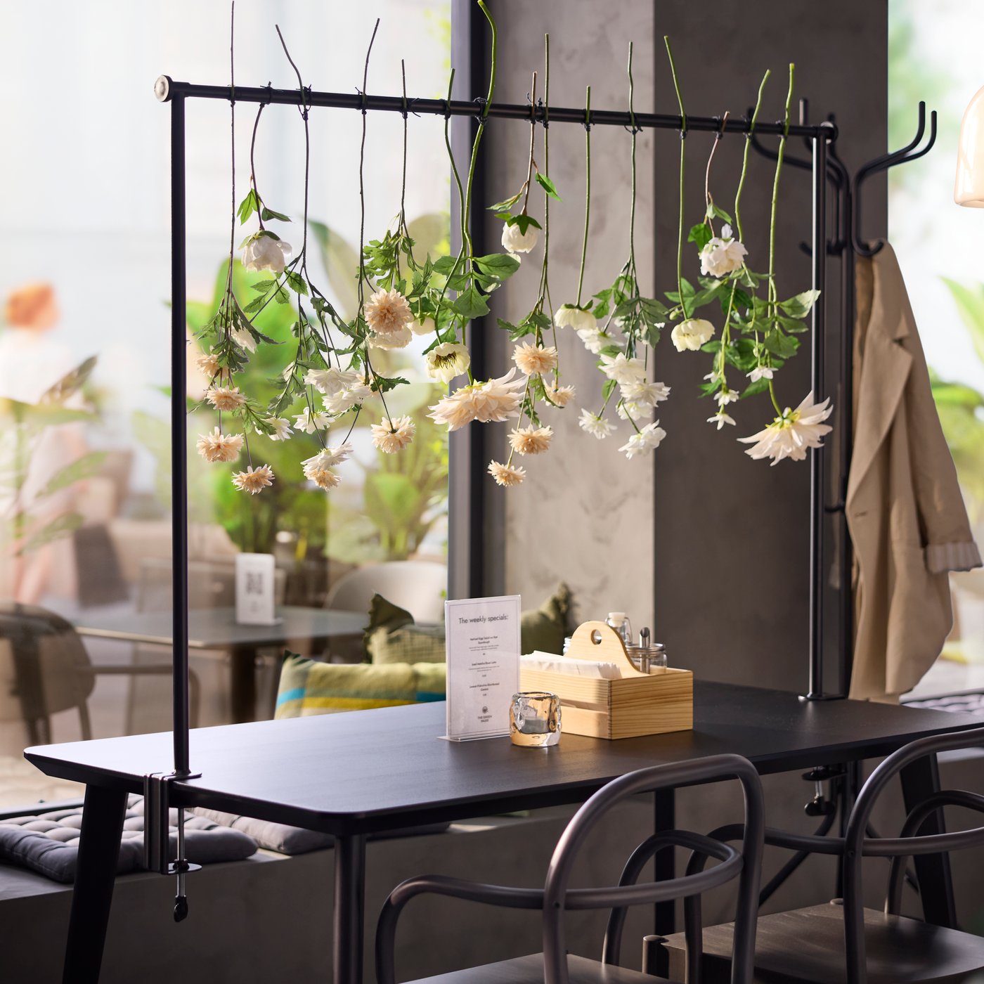 A seating area featuring a black HELGEÖ decorating rod for a table with hanging flowers and greenery in a bright café scene.