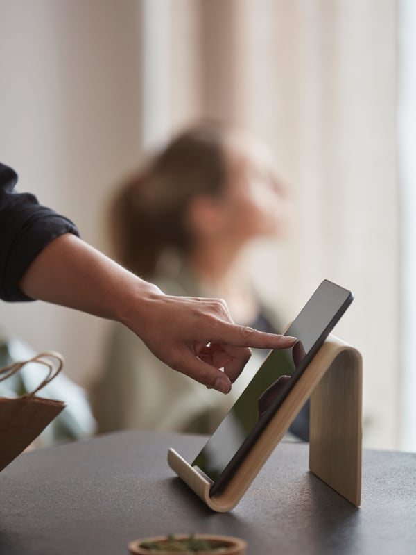 A hand is using an index finger to touch the display of a black tablet that is placed on a wooden stand on a dark grey table.