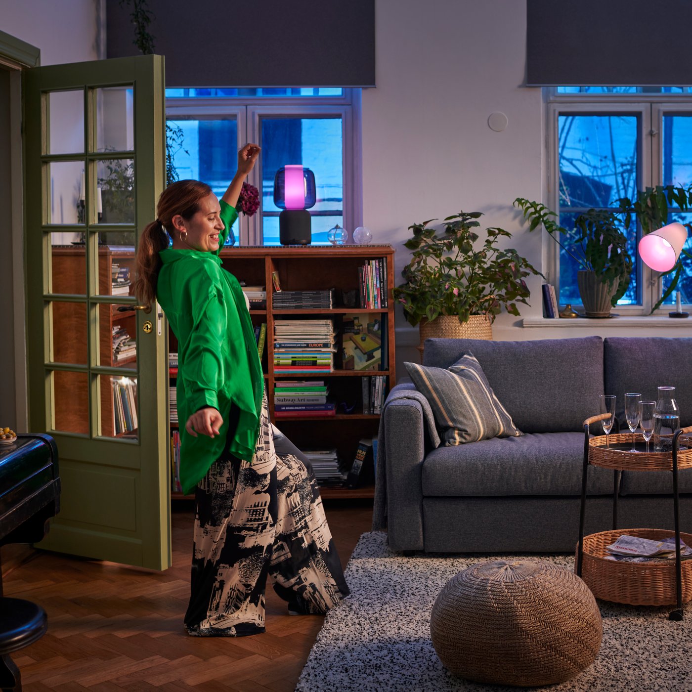 Una signora con un vestito verde sta ballando in un salotto con un tappeto bianco VINDUM e un divano grigio VIMLE.