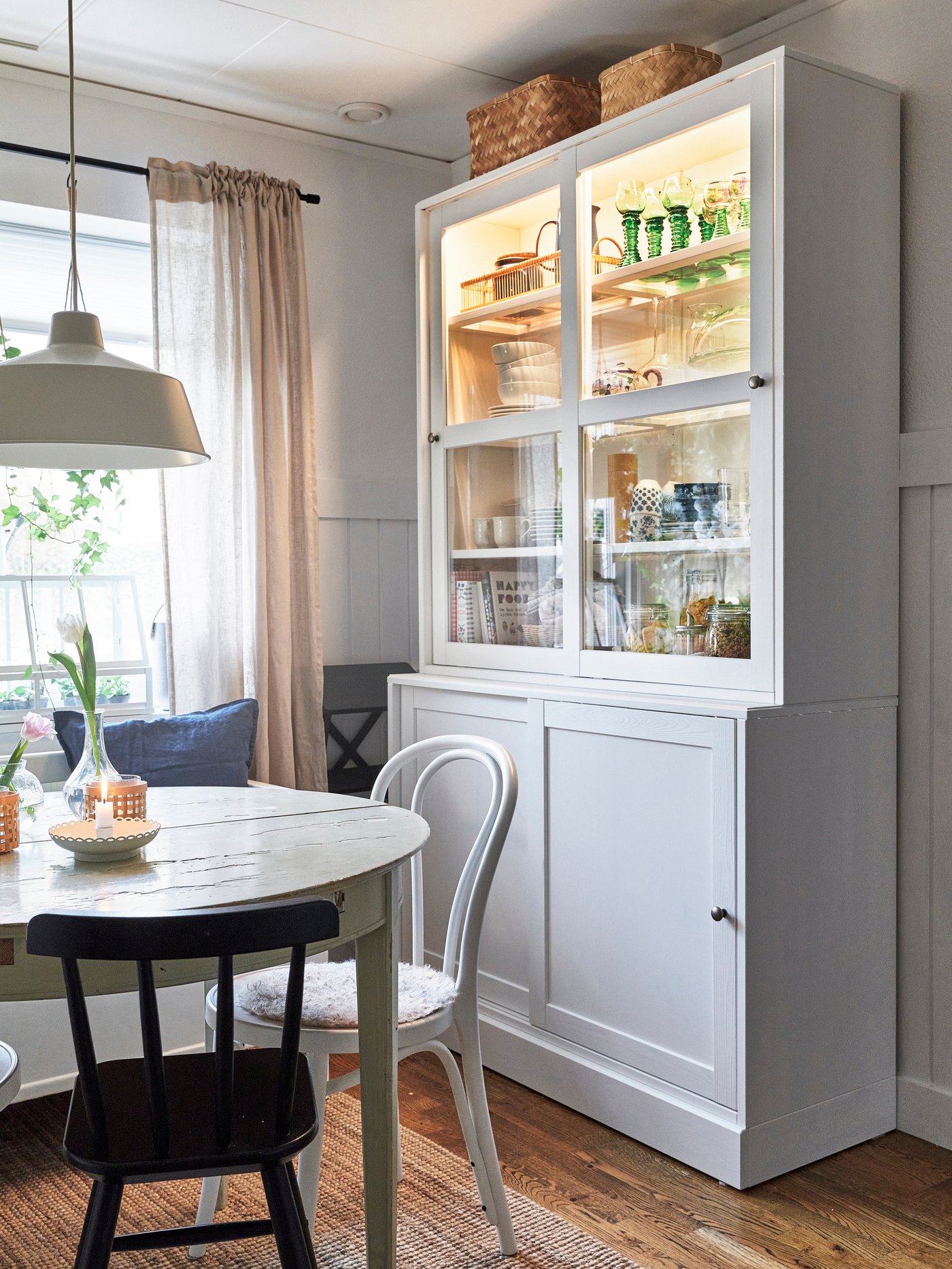 Matplats med runt vitt bord och stolar framför ett vitt vitrinskåp med glasdörrar. Skåpet är upplyst inifrån och visar glas och porslin.