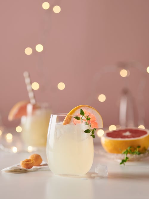 Într-o sufragerie există un pahar STORSINT transparent, umplut cu o băutură galbenă și garnisit cu o bucată de grapefruit.
