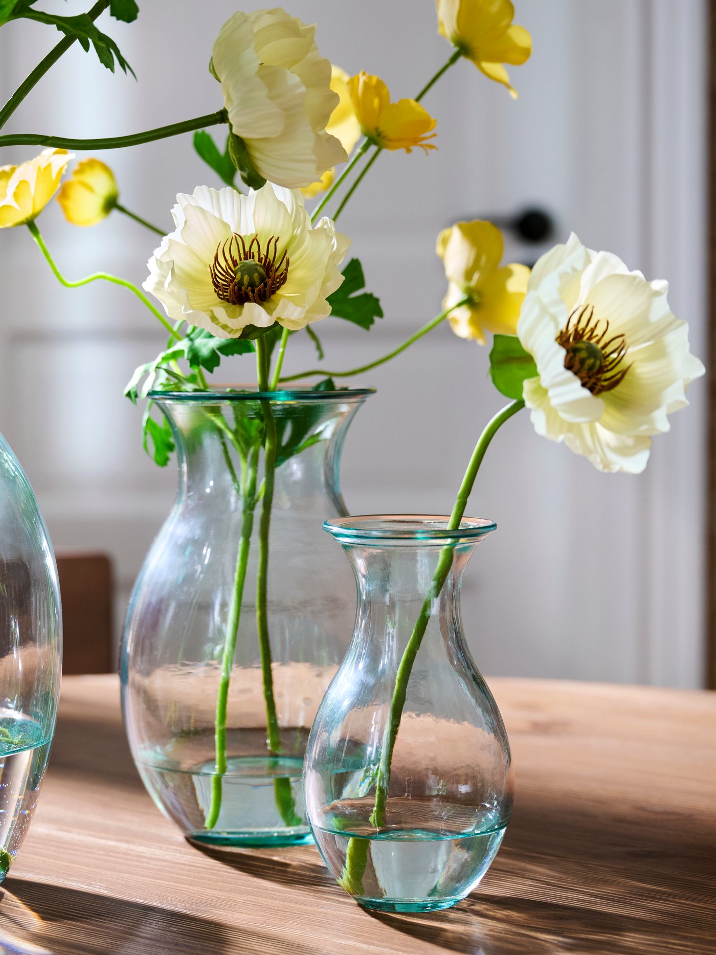 Three BERGKÖRSBÄR vases in clear glass, placed on a wooden table, each holding water and yellow flowers with green stems.
