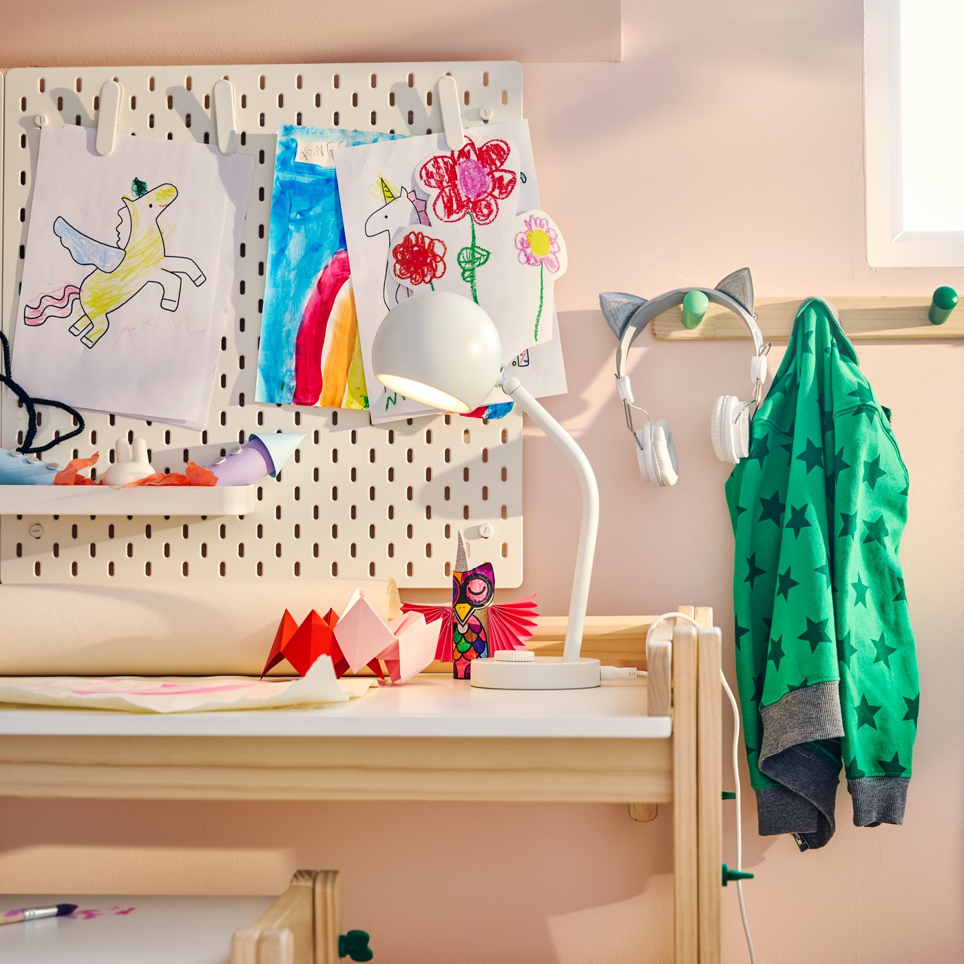 A pink children’s room with a white KORVSNÖRE lamp on a pine desk, beside a pegboard wall displaying children’s artwork.